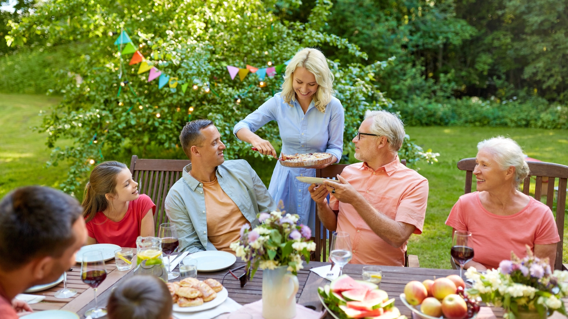 A multi-generational family gathering outdoors, enjoying a meal together in a lush garden setting. The scene features a woman serving food to an elderly man, with other family members smiling and engaged in conversation. The table is decorated with fresh fruits, flowers, and various dishes, emphasizing a warm, joyful atmosphere during a summer celebration. Perfect for SEO related to family gatherings, outdoor dining, and summer events.